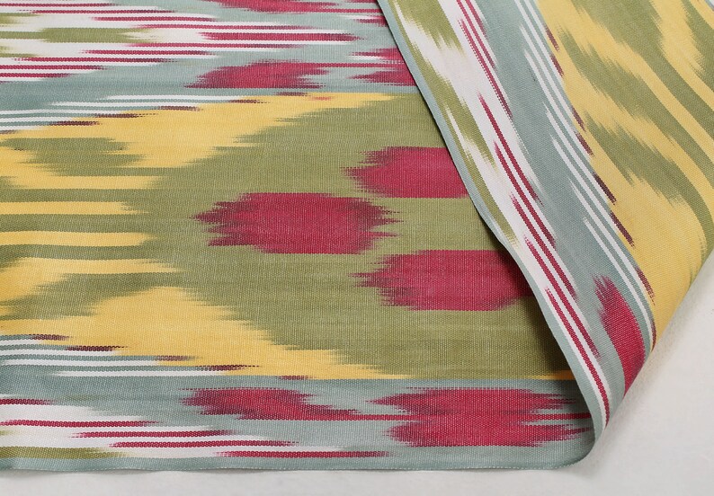 May include: A close-up of a fabric with a colorful ikat pattern. The fabric is a blend of yellow, green, red, and white. The pattern is a traditional ikat design, with a repeating motif of stripes and dots.