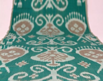 Könnte beinhalten: Nahaufnahme eines türkisfarbenen Stoffes mit einem weißen und braunen Ikat-Muster. Der Stoff hat eine gewebte Textur und einen dezenten Glanz.