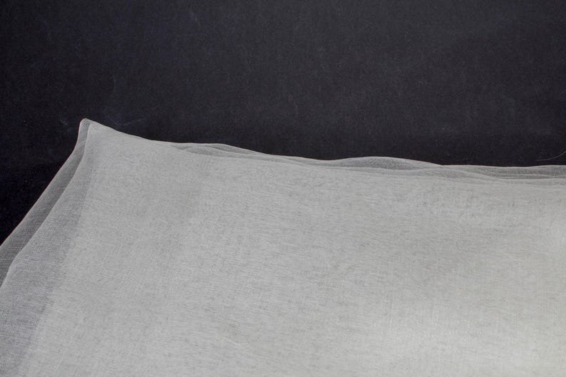 May include: A close-up of a white, sheer fabric with a delicate texture. The fabric is draped, creating soft folds and a subtle, undulating edge. The background is a dark, solid color.