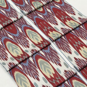 Pu&ograve; includere: Due pezzi di tessuto con un motivo ikat rosso, blu e crema. Il tessuto &egrave; piegato e impilato su una superficie bianca.