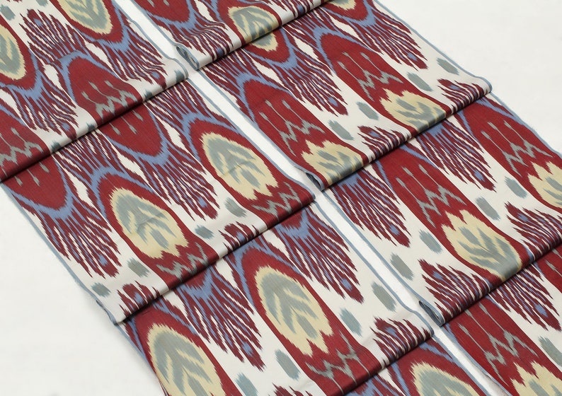 Pu&ograve; includere: Due pezzi di tessuto con un motivo ikat rosso, blu e crema. Il tessuto &egrave; piegato e impilato su una superficie bianca.