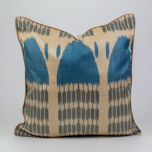 Puede incluir: Cojín decorativo cuadrado con un estampado ikat azul y beige. El cojín tiene un ribete marrón y un fondo neutro. El patrón presenta líneas verticales y formas de arco en azul y gris.