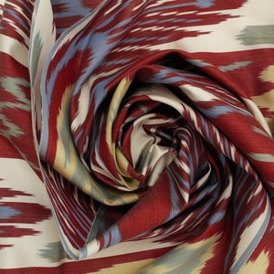 Pu&ograve; includere: Un primo piano di un tessuto a motivi rossi, blu e crema con un design ikat. Il tessuto &egrave; piegato e attorcigliato a forma di spirale.