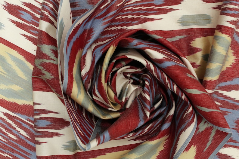 Pu&ograve; includere: Un primo piano di un tessuto a motivi rossi, blu e crema con un design ikat. Il tessuto &egrave; piegato e attorcigliato a forma di spirale.