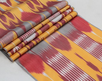 May include: A close-up of a fabric with a yellow, red, white, and gray ikat pattern. The fabric is folded in several layers, showing the pattern from different angles.