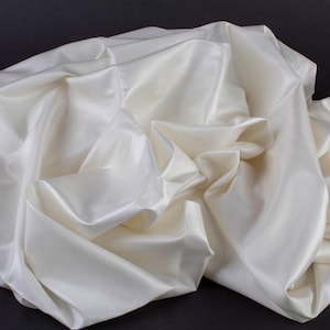 Könnte beinhalten: Nahaufnahme eines großen Stücks elfenbeinfarbenen Satin-Stoffs. Der Stoff ist drapiert und gefaltet, wodurch weiche Schatten und Glanzlichter entstehen. Das Material hat eine glatte, glänzende Oberfläche, die auf eine luxuriöse Qualität hindeutet. Der Hintergrund ist dunkel.