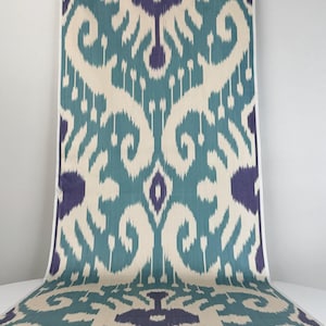 May include: A long strip of fabric featuring a repeating ikat pattern in teal, deep purple, and off-white. The design consists of large, symmetrical, abstract floral-like motifs with soft, blurred edges characteristic of the ikat dyeing technique.