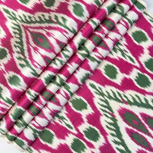 May include: Folded fabric with a vibrant ikat pattern. The design features a repeating motif of magenta, white, and green shapes. The fabric is folded, showcasing the pattern's intricate details and color variations. The textile appears to be silk or a similar luxurious material.