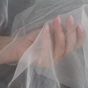 以下が含まれることがあります： 繊細な織り目を持つ、透明感のある白い生地のクローズアップ。生地はドレープされており、柔らかい折り目と影を作り出しています。この画像は、素材の透明感と軽さを強調しています。