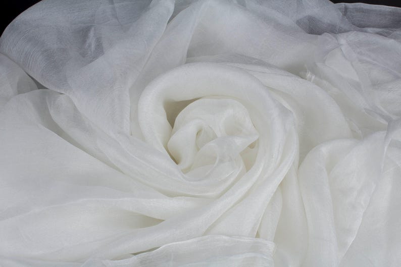 May include: A close-up shot of white fabric, possibly silk or organza, with a soft, flowing texture. The fabric is arranged in swirling folds, creating a sense of depth and movement. The color is a bright, clean white.