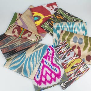 May include: A collection of folded silk ikat fabric pieces in various colors and patterns. The fabrics display a range of hues, including red, blue, green, and pink, with intricate, abstract designs. The pieces are arranged in a circular pattern.