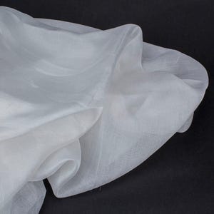May include: A close-up of a white, sheer fabric with a flowing, draped appearance. The fabric has a soft texture and is slightly translucent, with visible folds and shadows. The background is black.