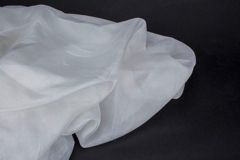 May include: A close-up of a white, sheer fabric with a flowing, draped appearance. The fabric has a soft texture and is slightly translucent, with visible folds and shadows. The background is black.