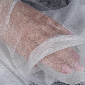 K&ouml;nnte beinhalten: Nahaufnahme eines transparenten wei&szlig;en Stoffes mit einem zarten, offenen Gewebe. Der Stoff ist drapiert und gefaltet, wodurch weiche Schatten und Lichteffekte entstehen. Die Textur wirkt leicht und luftig, was auf einen Textil f&uuml;r Kleidung oder Heimdekoration hindeutet.