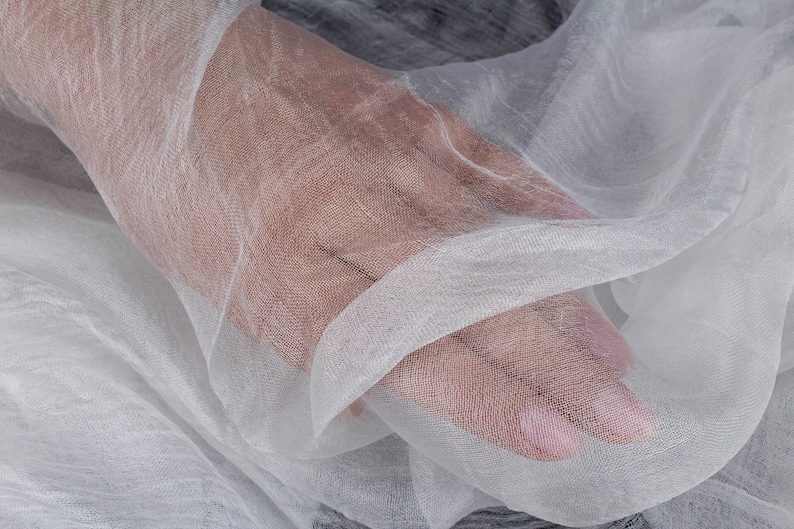 K&ouml;nnte beinhalten: Nahaufnahme eines transparenten wei&szlig;en Stoffes mit einem zarten, offenen Gewebe. Der Stoff ist drapiert und gefaltet, wodurch weiche Schatten und Lichteffekte entstehen. Die Textur wirkt leicht und luftig, was auf einen Textil f&uuml;r Kleidung oder Heimdekoration hindeutet.