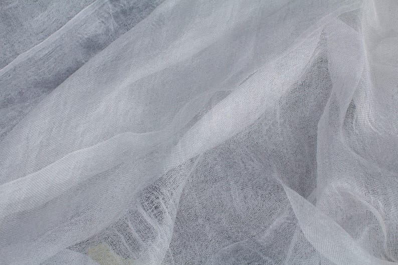K&ouml;nnte beinhalten: Nahaufnahme eines wei&szlig;en, transparenten Stoffes mit zarter, gewebter Textur. Der Stoff ist drapiert und gefaltet, wodurch weiche Schatten und Lichtreflexe entstehen. Das Material wirkt leicht und luftig.