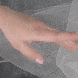 Pode incluir: Uma mão com unhas cuidadas repousa sobre um tecido transparente e claro. O tecido parece ser uma tecelagem delicada, com uma textura subtil. A imagem está bem iluminada, destacando os detalhes do tecido e da mão.