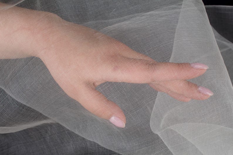 May include: A hand with manicured nails rests on a sheer, light-colored fabric. The fabric appears to be a delicate weave, with a subtle texture. The image is well-lit, highlighting the details of the fabric and the hand.