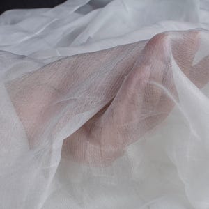 May include: Close-up of sheer white fabric, possibly chiffon or silk, draped and held. The fabric is translucent, allowing the skin of a hand to be partially visible through the delicate material. The folds create shadows and texture.