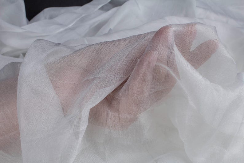 May include: Close-up of sheer white fabric, possibly chiffon or silk, draped and held. The fabric is translucent, allowing the skin of a hand to be partially visible through the delicate material. The folds create shadows and texture.