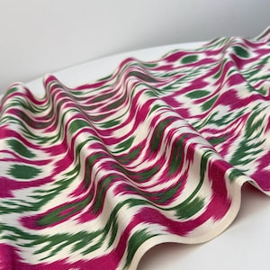 May include: A flowing fabric with a repeating pattern of magenta and green brushstrokes on an off-white background. The fabric is draped, showing the texture and design. The edges are finished with a cream-colored border.