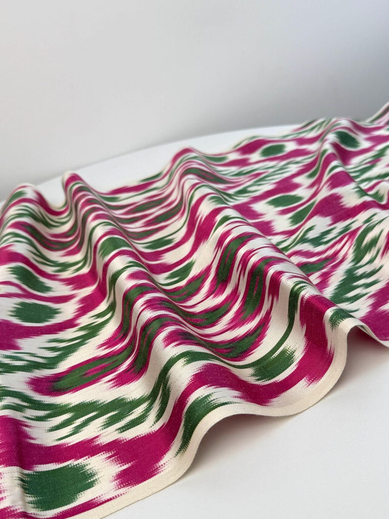 May include: A flowing fabric with a repeating pattern of magenta and green brushstrokes on an off-white background. The fabric is draped, showing the texture and design. The edges are finished with a cream-colored border.