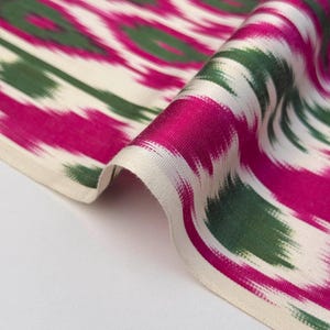May include: Close-up of a folded fabric with a vibrant ikat pattern. The design features bold fuchsia, green, and white brushstrokes creating an abstract, flowing motif. The fabric's texture is visible, suggesting a soft, possibly silk, material.