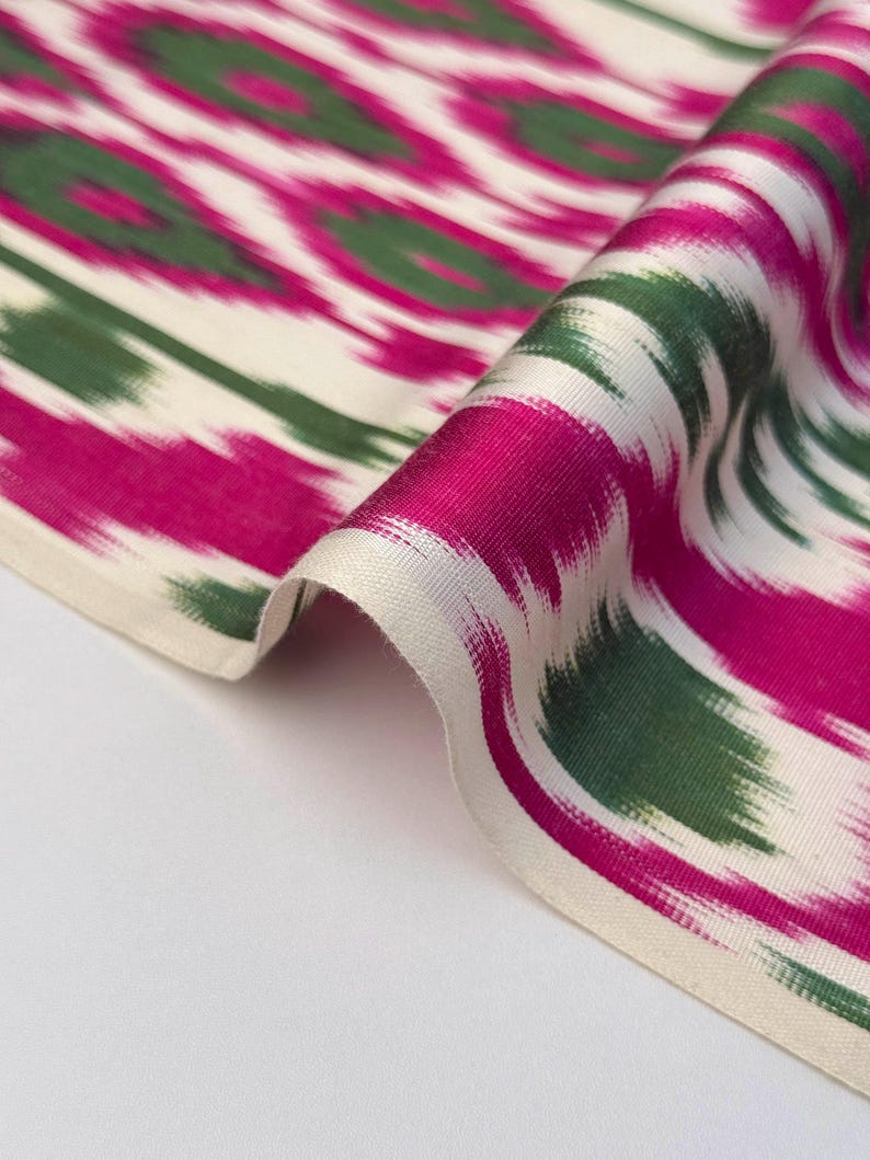 May include: Close-up of a folded fabric with a vibrant ikat pattern. The design features bold fuchsia, green, and white brushstrokes creating an abstract, flowing motif. The fabric's texture is visible, suggesting a soft, possibly silk, material.