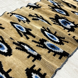 Op de afbeelding: Een gevouwen textiel met een herhalend patroon van zwarte, blauwe en witte oogvormige vormen op een beige achtergrond. De stof heeft een geribbelde textuur, waardoor een visuele diepte ontstaat. Het ontwerp is symmetrisch en visueel opvallend.