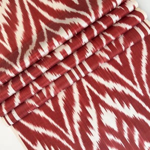 May include: A length of fabric featuring a bold ikat pattern in deep red and off-white. The textile displays a repeating chevron-like design with soft, blurred edges characteristic of the ikat dyeing technique, folded to show multiple layers.