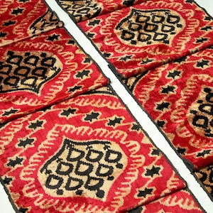 May include: Two strips of red velvet fabric with a black and gold ikat pattern. The fabric has a raised texture and a luxurious feel.
