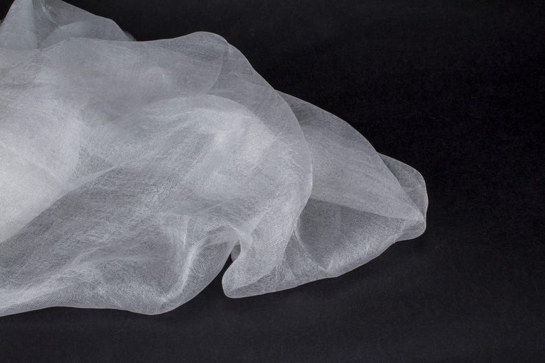 K&ouml;nnte beinhalten: Nahaufnahme eines wei&szlig;en, durchscheinenden Stoffes mit zarter, luftiger Textur. Der Stoff ist drapiert und gefaltet, wodurch weiche Schatten vor einem dunklen Hintergrund entstehen. Das Material wirkt leicht und flie&szlig;end.
