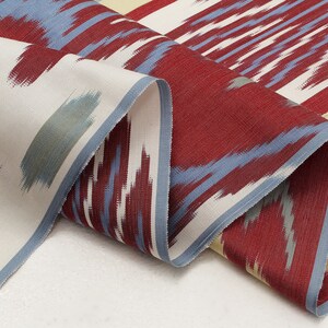 Pu&ograve; includere: Tessuto a motivi rossi, blu e crema con un design ikat. Il tessuto &egrave; piegato e drappeggiato su una superficie bianca.