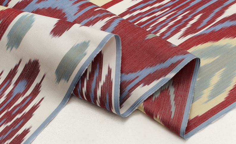 Pu&ograve; includere: Tessuto a motivi rossi, blu e crema con un design ikat. Il tessuto &egrave; piegato e drappeggiato su una superficie bianca.