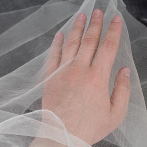 May include: Close-up of sheer, white fabric with a delicate weave. The fabric is draped, creating soft folds and shadows. The texture appears lightweight and airy, suitable for apparel or decorative purposes.