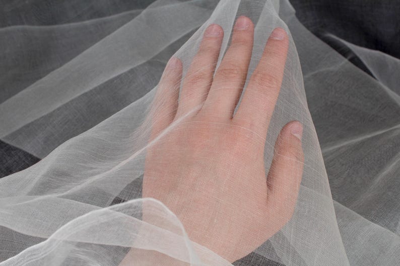 May include: Close-up of sheer, white fabric with a delicate weave. The fabric is draped, creating soft folds and shadows. The texture appears lightweight and airy, suitable for apparel or decorative purposes.