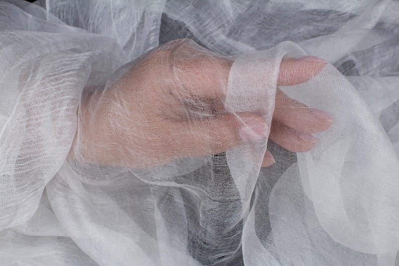 K&ouml;nnte beinhalten: Nahaufnahme eines durchsichtigen wei&szlig;en Stoffes, m&ouml;glicherweise Gaze oder Musselin, drapiert und gehalten. Der Stoff ist durchscheinend, so dass die Haut einer Hand teilweise durch das Material sichtbar ist. Die Textur wirkt weich und zart.
