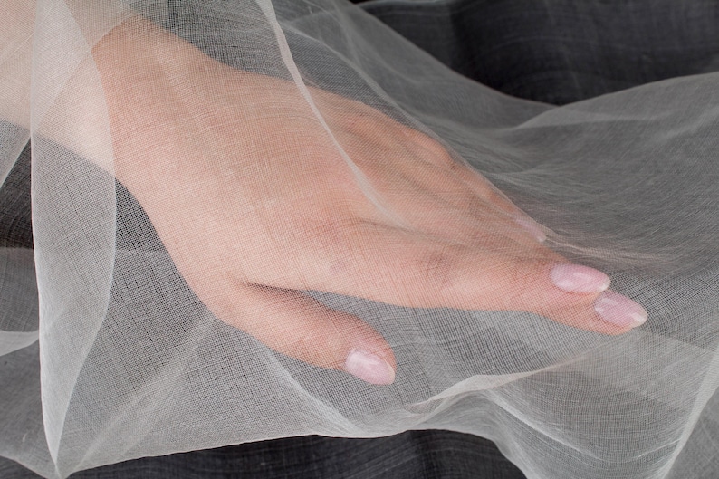 May include: Close-up of a hand resting on a sheer, white fabric. The fabric is draped, creating soft folds and shadows. The hand has pale skin and polished nails. The background is a darker, textured material.