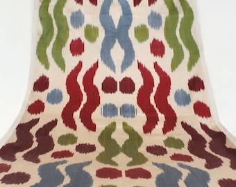 May include: A beige fabric with a repeating pattern of red, green, blue, and brown abstract shapes. The fabric has a subtle sheen and a soft texture.