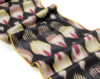 May include: A black silk fabric with a colourful ikat pattern. The fabric has a yellow border and is folded in a wavy pattern.