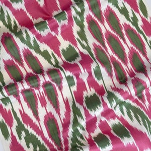 May include: A length of fabric featuring a vibrant ikat pattern with elongated, abstract shapes in shades of magenta, deep forest green, and crisp white. The design consists of repeating vertical motifs with soft, blurred edges throughout.