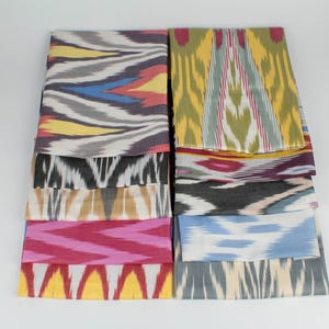 May include: A collection of folded fabric samples with vibrant ikat patterns. The textiles feature a variety of colors, including yellow, pink, blue, gray, and white. The designs are geometric and abstract, suitable for home decor or fashion.