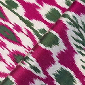 May include: Close-up of a folded fabric with a vibrant ikat pattern. The design features diagonal stripes in fuchsia, green, and white. The fabric has a silky sheen, suggesting a luxurious material suitable for apparel or home decor.