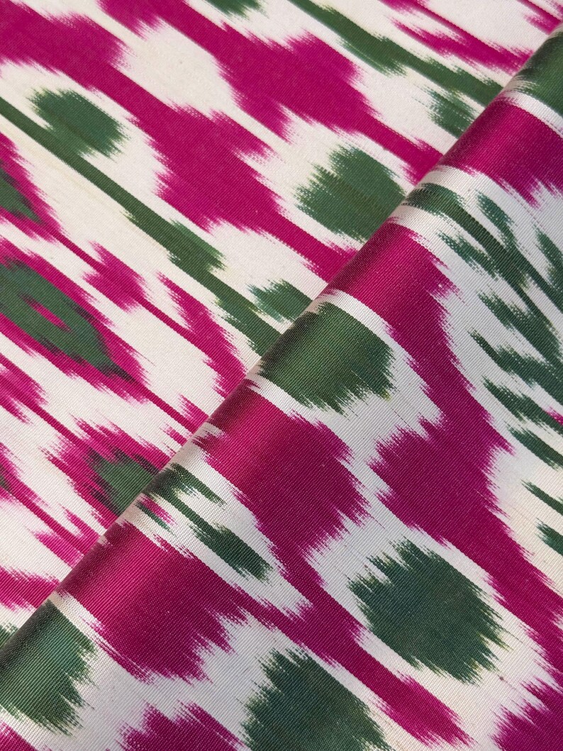 May include: Close-up of a folded fabric with a vibrant ikat pattern. The design features diagonal stripes in fuchsia, green, and white. The fabric has a silky sheen, suggesting a luxurious material suitable for apparel or home decor.