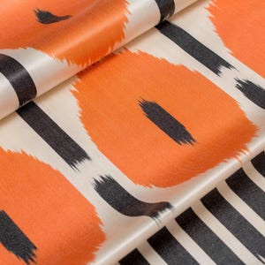 Op de afbeelding: Close-up van een gevouwen stof met een herhalend patroon. Het ontwerp heeft grote oranje vormen met zwarte penseelstreken en zwarte verticale strepen op een gebroken witte achtergrond. De stof heeft een satijnen glans.