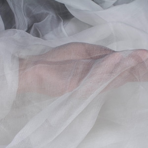 May include: Close-up of sheer white fabric, possibly silk or chiffon, draped and flowing. The fabric is translucent, with a hand visible beneath the layers. The material has a delicate texture and appears soft. Ideal for apparel or craft projects.