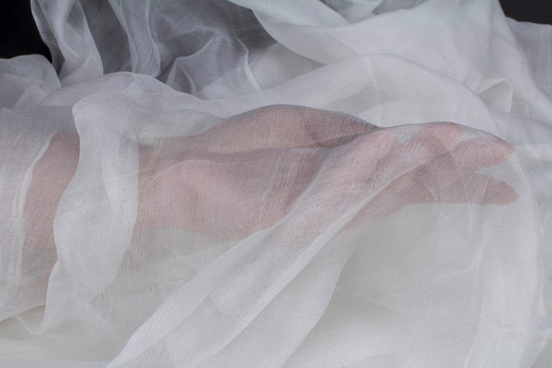 May include: Close-up of sheer white fabric, possibly silk or chiffon, draped and flowing. The fabric is translucent, with a hand visible beneath the layers. The material has a delicate texture and appears soft. Ideal for apparel or craft projects.