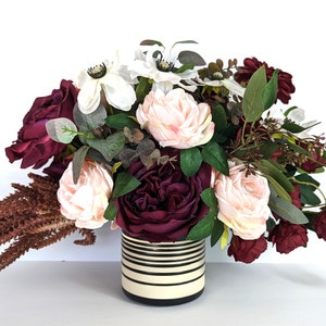 May include: A floral arrangement in a black and white striped vase. The arrangement features pink, white, and burgundy roses, along with greenery.