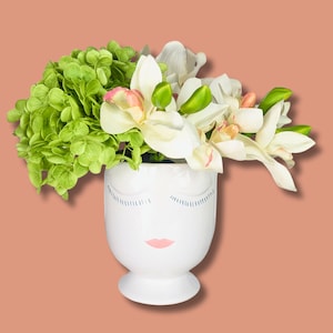 May include: White ceramic vase with a stylized face design, featuring closed eyes and pink lips. The vase is filled with a bouquet of white orchids and green hydrangeas.