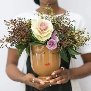 May include: A brown ceramic vase with a stylized face design, featuring closed eyes and a red mouth, holds a bouquet of white and purple flowers and brown foliage.